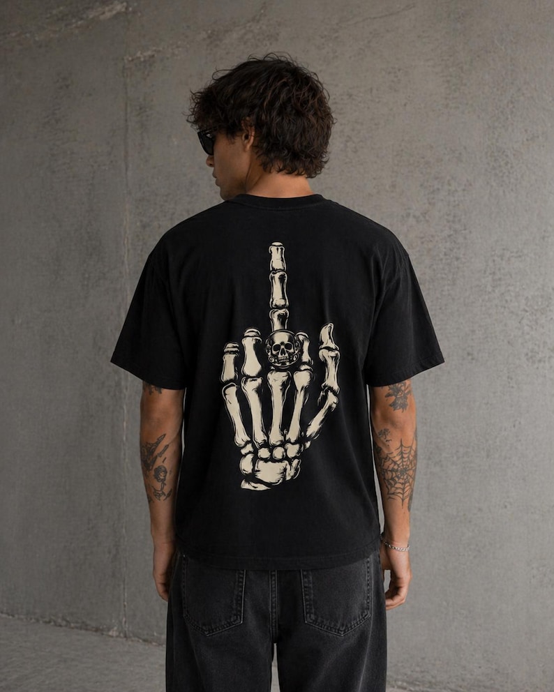 May include: Black t-shirt featuring a skeletal hand giving the middle finger. The graphic is a detailed illustration of a bone hand with a skull on the middle finger. The shirt is a dark color and has a casual fit.