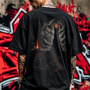 Könnte beinhalten: Ein schwarzes T-Shirt mit kurzen Ärmeln und einem detaillierten Grafikdruck auf der Rückseite. Das Motiv zeigt eine dunkle, hoch aufragende Festung mit einem leuchtenden orangefarbenen Auge an der Spitze, einen Vulkanausbruch und zwei kleine Figuren.
