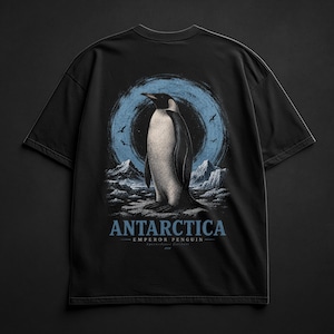 May include: A black short-sleeve t-shirt featuring a graphic print of an emperor penguin standing on ice against a blue circular background. The text "ANTARCTICA" and "EMPEROR PENGUIN" is printed in blue below the central penguin illustration.