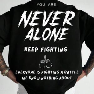 May include: Black sweatshirt with white text that reads "YOU ARE NEVER ALONE KEEP FIGHTING". Below the text are two boxing gloves. The bottom of the shirt reads "EVERYONE IS FIGHTING A BATTLE WE KNOW NOTHING ABOUT".