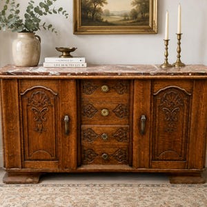 Könnte beinhalten: Ein hölzernes Sideboard mit einer braunen Marmorplatte, zwei Seitenschränken mit geschnitzten floralen Details und vier mittigen Schubladen mit kleinen runden Knöpfen. Die Schranktüren haben vertikale Metallgriffe und das Holz ist gemasert.