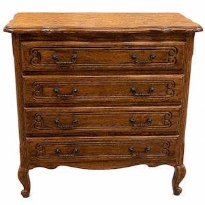 May include: A medium brown wooden chest of drawers featuring four horizontal drawers with carved scrollwork detailing on the drawer fronts. The piece stands on four curved cabriole legs and includes dark metal hardware pulls on each drawer.