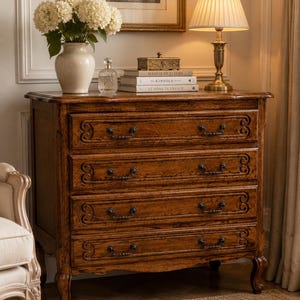 May include: A wooden chest of drawers with four horizontal drawers, each featuring dark metal handles and carved scrollwork details. The piece stands on four curved legs and is topped with a white vase of flowers, a glass bottle, and books.