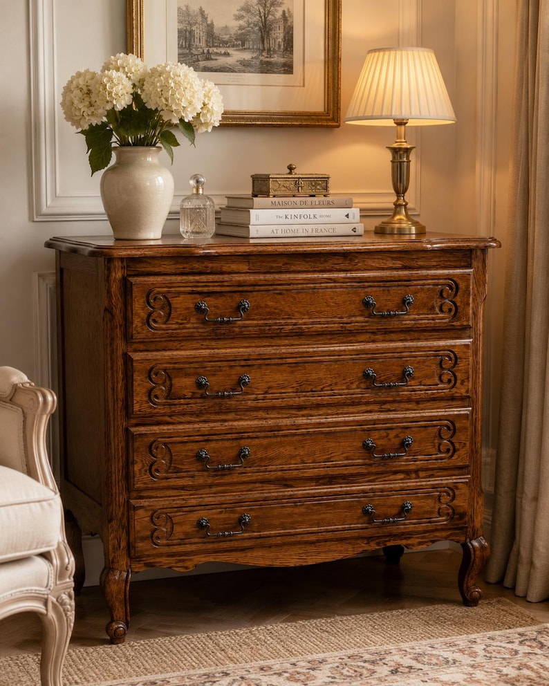 May include: A wooden chest of drawers with four horizontal drawers, each featuring dark metal handles and carved scrollwork details. The piece stands on four curved legs and is topped with a white vase of flowers, a glass bottle, and books.