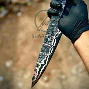 May include: A handcrafted knife with a patterned blade, held by a gloved hand. The blade features a unique design with black, white, and copper tones. The knife's handle is not visible. The image is set against a blurred, neutral background.