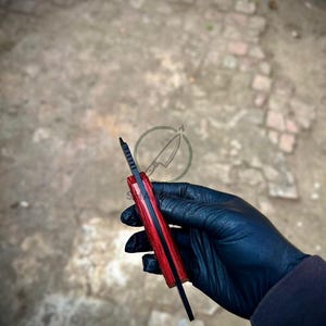 May include: A fixed-blade knife featuring a black metal spine with serrated jimping and a handle constructed from two vibrant red wood scales. The knife is held vertically, showcasing the contrast between the dark blade and the bright red handle.