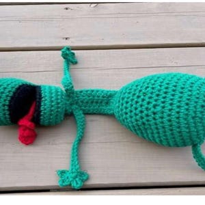May include: A handmade green crochet frog toy with white eyes, a black mouth, and a red tongue. The frog is lying on its back with its arms and legs outstretched. The toy is made of soft yarn.