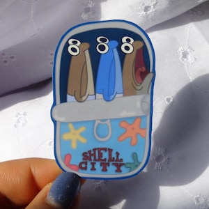 May include: A blue and white sticker with a sardine can design. The sticker features three cartoon fish in brown, blue, and tan. The text "888" is above the fish, and "SHELL CITY" is written below the fish.