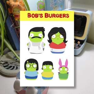 May include: A white card with the text "BOB'S BURGERS" in red and yellow. The card features cartoon illustrations of the Belcher family: Bob, Linda, Tina, Gene, and Louise. A blue retro-style television is in the background.