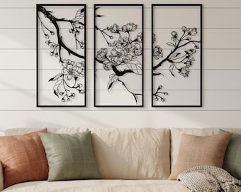 May include: Three framed black metal wall art pieces depicting cherry blossom branches. The artwork is mounted on a white horizontal plank wall above a beige sofa with various throw pillows. A woven basket and a lamp are in the foreground.