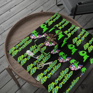 Op de afbeelding: Zwart cadeaupapier met een patroon van groene aliens op eenhoorns en de tekst "It's a alien" in neon groen en blauw. Een ingepakt cadeau met een bruine strik ligt op het papier, samen met een schaar en tape.