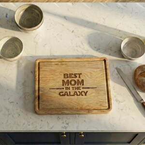 Può includere: Un tagliere in legno con la scritta incisa "BEST MOM IN THE GALAXY". La tavola è rettangolare con un bordo rientrante. L'immagine mostra anche tre piccole ciotole, un coltello e un pane.