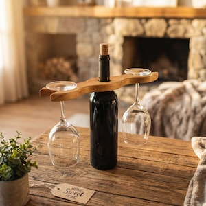 Puede incluir: Un soporte de madera para botellas de vino con dos copas de vino colgando. Una botella de vino oscuro con un corcho en el centro. Una etiqueta con las palabras "Home Sweet Home" está sobre la mesa.