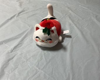 May include: A plush white cat toy wearing a Santa hat and red and white outfit. The cat has a red bow and green holly leaves on its hat. The toy is lying down on a light blue surface.