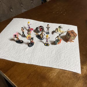 May include: A collection of small, colorful figurines, each depicting a unique character. The figurines are set on a white paper towel, with a wooden table in the background. The characters have various poses and outfits.