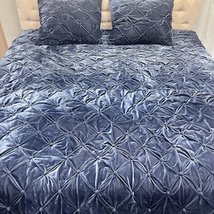 Op de afbeelding: Een marineblauwe fluwelen beddenset met een quilt en twee bijpassende vierkante kussens. De quilt en kussens hebben een gewatteerd ruitpatroon. Het hoofdbord is lichtbeige.