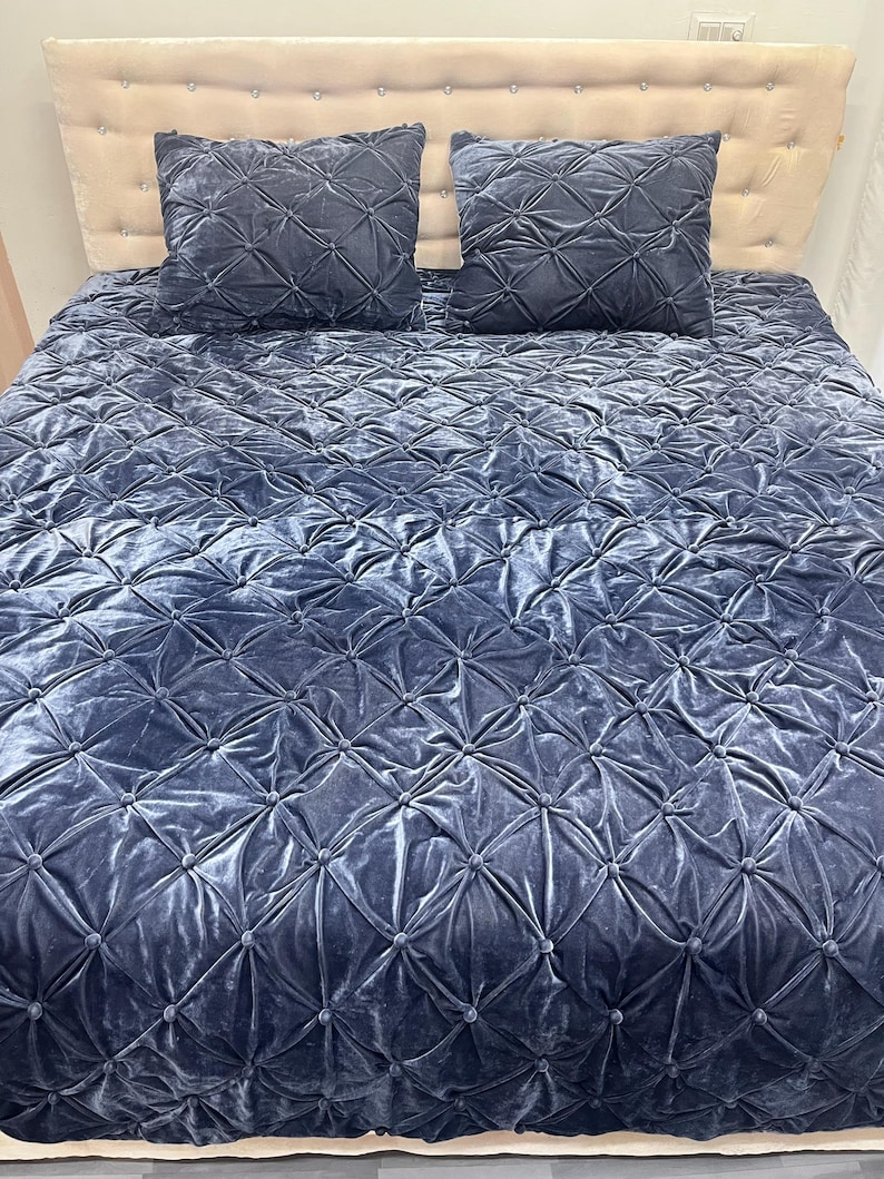 Op de afbeelding: Een marineblauwe fluwelen beddenset met een quilt en twee bijpassende vierkante kussens. De quilt en kussens hebben een gewatteerd ruitpatroon. Het hoofdbord is lichtbeige.