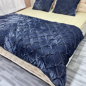 Op de afbeelding: Een marineblauwe fluwelen sprei en twee bijpassende kussens met een geometrisch gecapitonneerd ontwerp. De sprei bedekt een bed met een cr&egrave;mekleurige matras en hoofdbord. De kussens zijn vierkant en hebben een vergelijkbaar gecapitonneerd patroon.