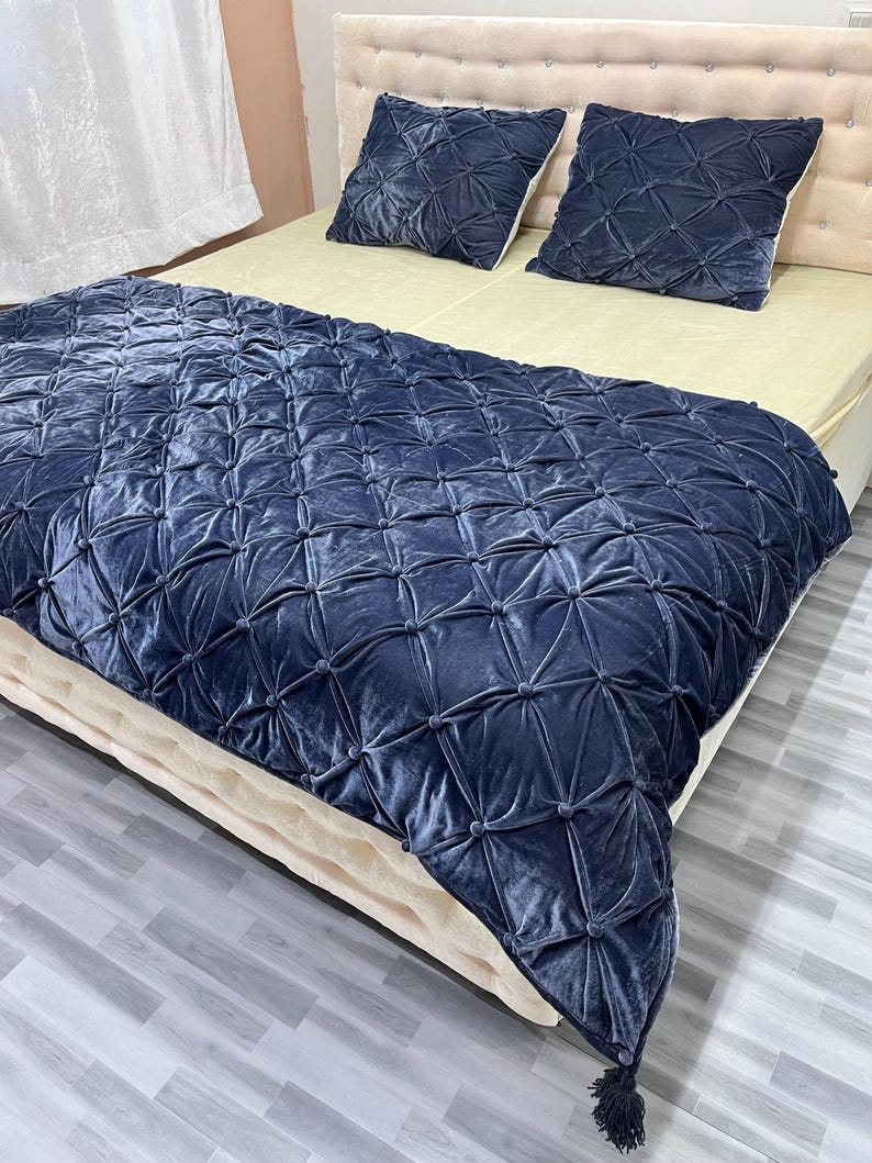 Op de afbeelding: Een marineblauwe fluwelen sprei en twee bijpassende kussens met een geometrisch gecapitonneerd ontwerp. De sprei bedekt een bed met een cr&egrave;mekleurige matras en hoofdbord. De kussens zijn vierkant en hebben een vergelijkbaar gecapitonneerd patroon.