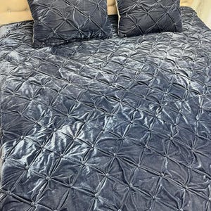 Op de afbeelding: Een beddenset met een gewatteerde, fluweelachtige sprei en twee bijpassende vierkante kussens. De set heeft een gedempte blauwgrijze kleur, met een ruitpatroon dat door de stiksels is gecre&euml;erd. Het hoofdbord is lichtbeige.