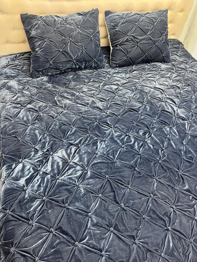 Op de afbeelding: Een beddenset met een gewatteerde, fluweelachtige sprei en twee bijpassende vierkante kussens. De set heeft een gedempte blauwgrijze kleur, met een ruitpatroon dat door de stiksels is gecre&euml;erd. Het hoofdbord is lichtbeige.