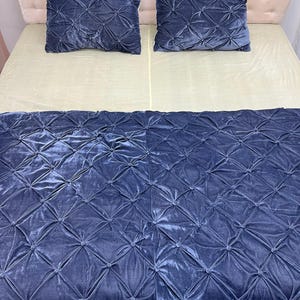 Op de afbeelding: Een bed met een lichtbeige matras en een marineblauwe fluwelen deken en twee bijpassende kussens. De deken en kussens hebben een ruitpatroon met knoopaccenten. Het hoofdbord is lichtroze.