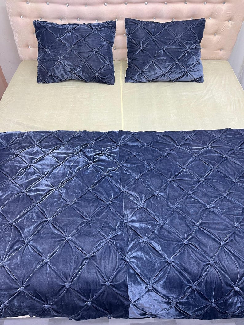 Op de afbeelding: Een bed met een lichtbeige matras en een marineblauwe fluwelen deken en twee bijpassende kussens. De deken en kussens hebben een ruitpatroon met knoopaccenten. Het hoofdbord is lichtroze.