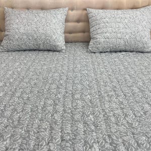 Könnte beinhalten: Ein grau-weißes Bettwäsche-Set mit floralem Muster und zwei passenden quadratischen Kissen. Die Bettwäsche weist ein sich wiederholendes botanisches Design auf und wird auf einem Bett mit beigem Kopfteil präsentiert.