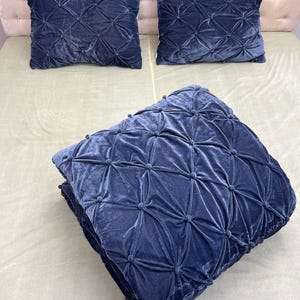 Op de afbeelding: Een set marineblauwe fluwelen beddengoed, bestaande uit twee vierkante kussens en een opgevouwen deken. De kussens en deken hebben een ruitvormig patroon met verhoogde, afgeronde accenten. De deken heeft een kwastjesrand.