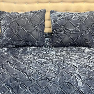 Op de afbeelding: Twee vierkante, donkergrijze fluwelen kussens met een diamantgestikt patroon liggen op een bijpassend sprei. De kussens en het sprei hebben een zachte, luxueuze uitstraling. Het hoofdbord is lichtbeige.