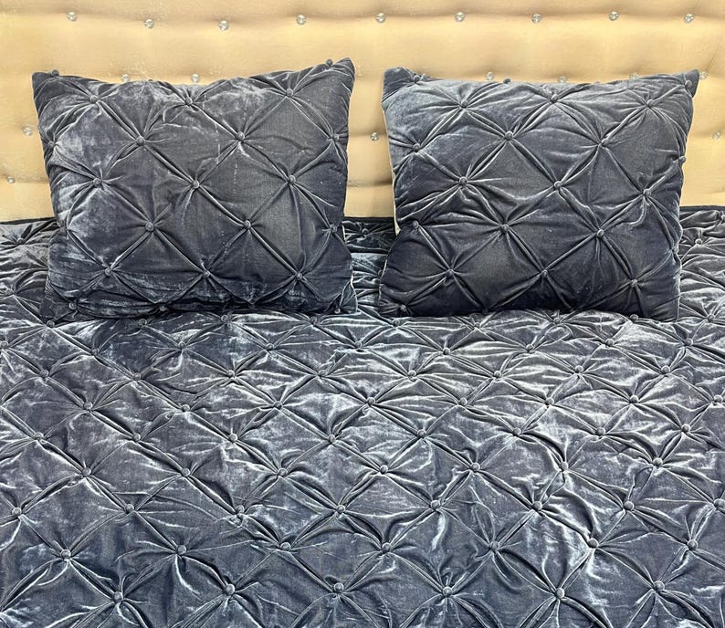 Op de afbeelding: Twee vierkante, donkergrijze fluwelen kussens met een diamantgestikt patroon liggen op een bijpassend sprei. De kussens en het sprei hebben een zachte, luxueuze uitstraling. Het hoofdbord is lichtbeige.