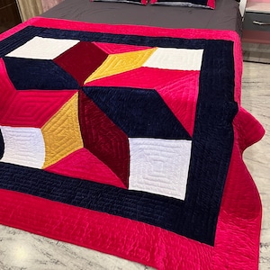 May include: A vibrant, geometric-patterned quilt and two matching pillow shams. The quilt features a patchwork design with red, white, navy, and yellow sections. The bedspread has a bright pink border and is displayed on a bed with a dark gray sheet.