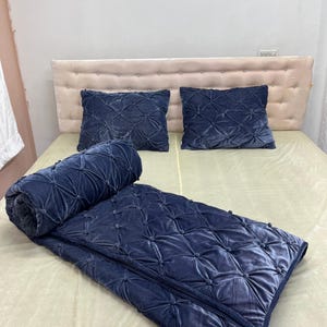 Op de afbeelding: Een marineblauwe fluwelen sprei en twee bijpassende kussens met een gewatteerd patroon. De sprei is aan &eacute;&eacute;n uiteinde opgerold en heeft een kwast. Het bed heeft een beige hoofdbord en een beige laken.