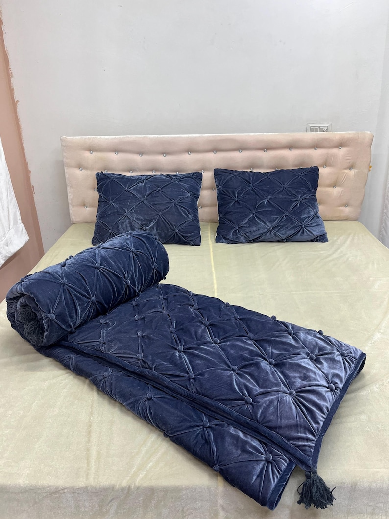 Op de afbeelding: Een marineblauwe fluwelen sprei en twee bijpassende kussens met een gewatteerd patroon. De sprei is aan &eacute;&eacute;n uiteinde opgerold en heeft een kwast. Het bed heeft een beige hoofdbord en een beige laken.