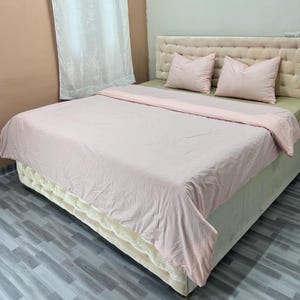 Könnte beinhalten: Ein Bett mit einer hellrosa Bettdecke und zwei passenden Kissen. Das Kopfteil ist hellbeige mit einem Knopf-getufteten Design. Der Bettrahmen ist hellbeige mit einem cremefarbenen Rock.