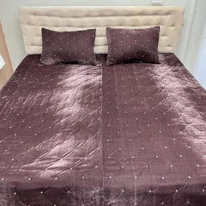 May include: A bed with a cream-colored headboard and a dark mauve velvet quilt. Two matching square pillows with a dotted pattern are on the bed. The quilt has a diamond pattern with small white dots.