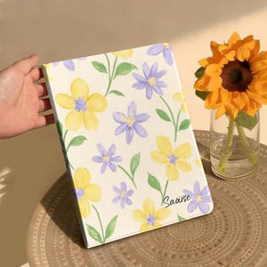 May include: A rectangular notebook with a cream-colored cover featuring a watercolor-style floral pattern of yellow and purple flowers with green stems and leaves. The name Saoirse is printed in a black script font on the lower right corner.
