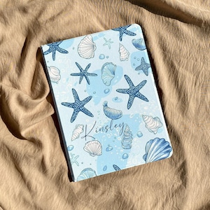 Op de afbeelding: Een rechthoekige tablethoes met een lichtblauwe achtergrond voorzien van een patroon van donkerblauwe zeesterren, diverse witte en blauwe schelpen en kleine belletjes. De naam Kinsley staat in het midden in een donkerblauw cursief lettertype.