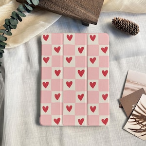Puede incluir: Una funda rosa para tableta con un patrón de tablero de ajedrez de cuadrados rosas y blancos, cada uno con un corazón rojo. La funda está cerrada y tiene una cubierta magnética.