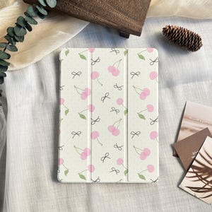 May include: A white tablet case with a repeating pattern of pink cherries, green leaves, and black bows. The case is on a cream-colored surface with a wooden box and pine cone in the background. The case is designed to protect a tablet.