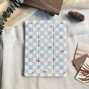 May include: A tablet case with a blue and white checkered pattern, adorned with red cherry illustrations. The case is designed to protect a tablet and features a folding cover. The overall aesthetic is a blend of functionality and a playful design.