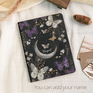 Pode incluir: Um diário preto com um design de borboletas e lua. A capa apresenta borboletas roxas, brancas e douradas, juntamente com uma lua crescente e o nome "Margaret". O texto "You can add your name" está na parte inferior.