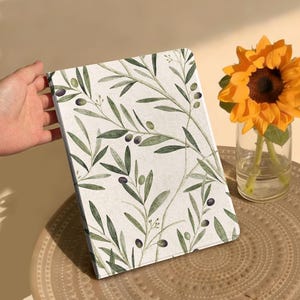Puede incluir: Un cuaderno con fondo blanco y un patrón repetido de ramas de olivo con hojas verdes y aceitunas negras. El cuaderno está sostenido por una mano. Un girasol en un jarrón de cristal está en el fondo.