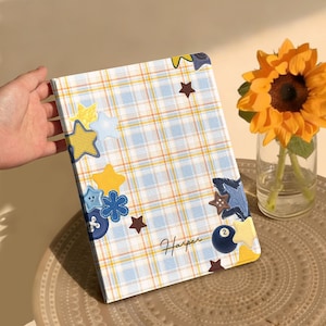 Puede incluir: Un cuaderno rectangular con una cubierta de cuadros en azul claro, blanco y amarillo. La cubierta está decorada con varios parches en forma de estrella, parches en forma de flor azul y una pequeña bola de billar negra con el número dos. El nombre Harper está impreso en letra cursiva cerca de la esquina inferior derecha.