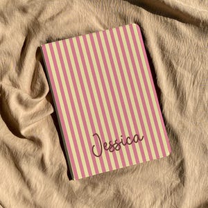 May include: A personalized notebook with a pink and cream striped pattern. The name "Jessica" is written in a cursive font on the cover. The notebook is rectangular and rests on a beige fabric surface.