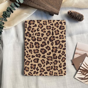 Può includere: Una custodia per tablet beige con un motivo leopardato marrone. La custodia rettangolare ha una copertura pieghevole. Lo sfondo include una scatola di legno, una pigna e alcune carte.