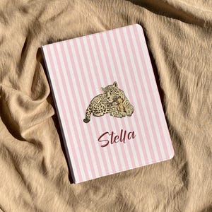 Puede incluir: Un cuaderno a rayas rosas y blancas con una ilustración de leopardo y el nombre "Stella". El leopardo es marrón con manchas negras. El cuaderno es rectangular.