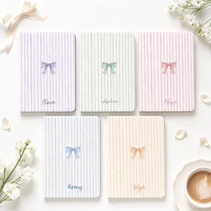 May include: Five rectangular notebooks with vertical striped covers in lavender, mint green, pink, light blue, and yellow. Each notebook features a centred watercolour-style bow illustration and a printed name: Claire, Archie, Hazel, Remy, and Maple.