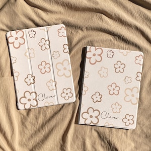 Op de afbeelding: Twee tablethoezen met een lichtbeige achtergrond en een herhalend patroon van omlijnde bloemen in bruin- en tinten. Elke hoes toont het woord Clover in een zwart cursief lettertype bij de rechterbenedenhoek.