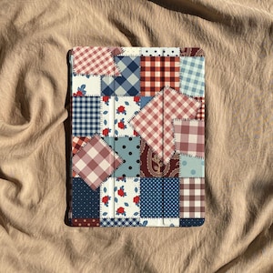 Könnte beinhalten: Eine farbenfrohe Tablet-Hülle mit Patchwork-Design. Die Hülle besteht aus Stoffquadraten in verschiedenen Mustern, darunter Karos, Tupfen und Blumendrucke. Die Farbpalette umfasst Rot, Blau und Weiß.